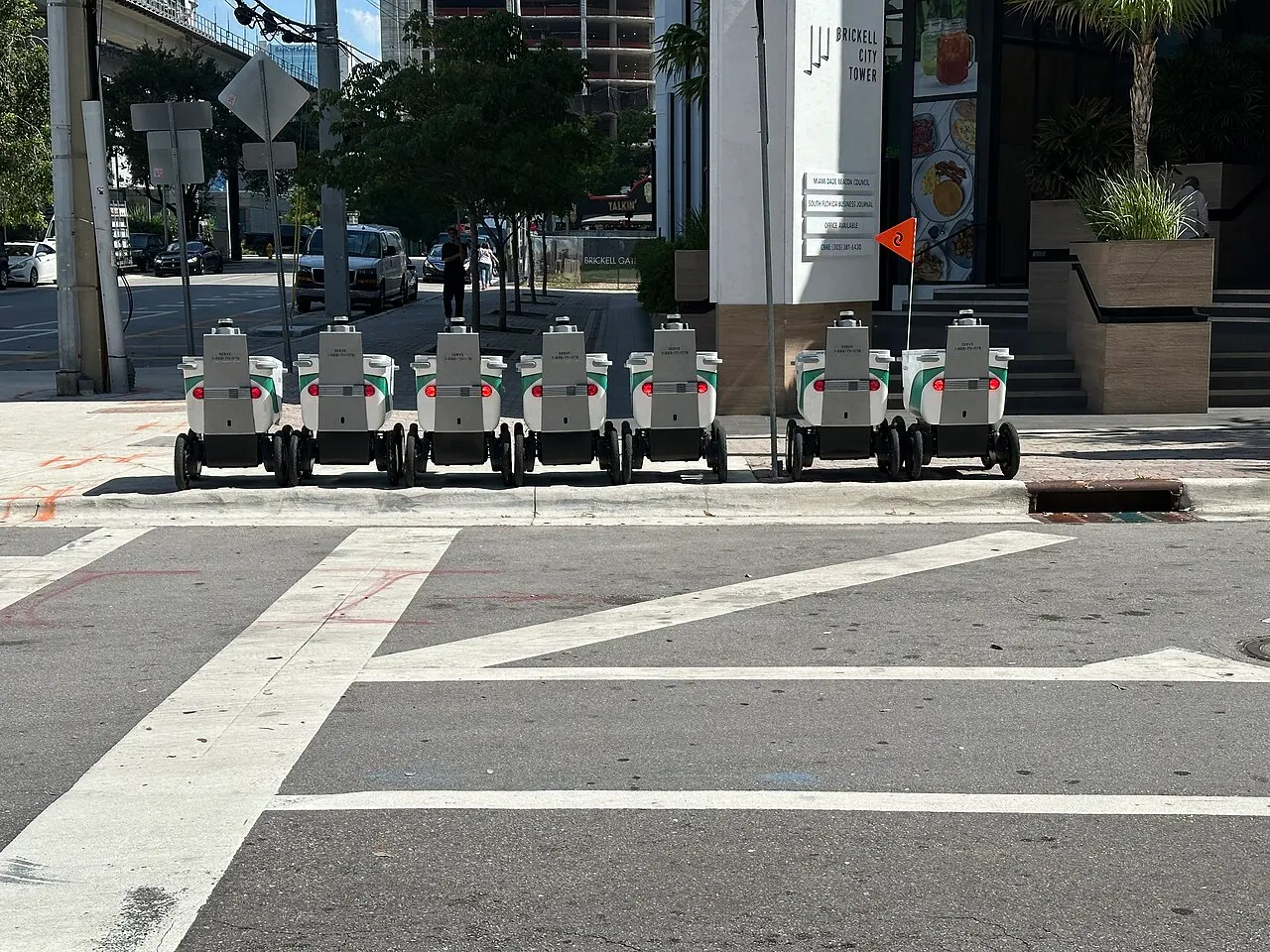 Illustrazione piatta di un pedone in piedi davanti a un piccolo robot per le consegne su ruote, su un marciapiede cittadino.